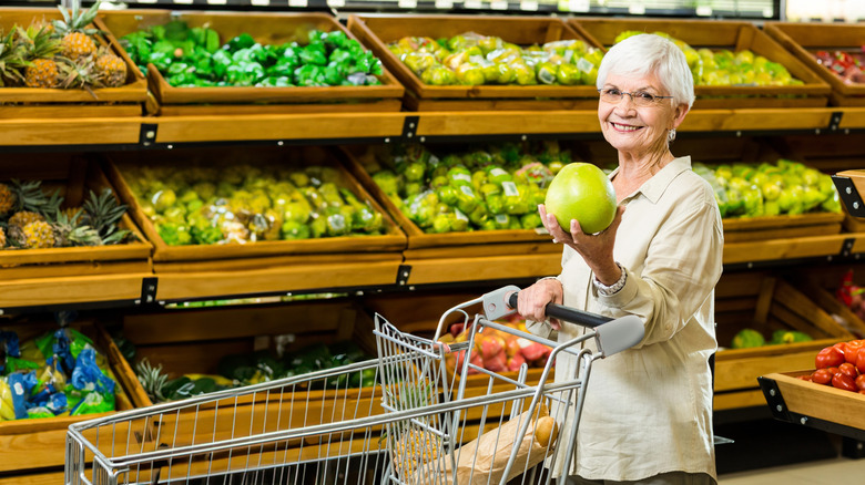 Senior shopper holding green apple while pushing metal cart in grocery produce aisle