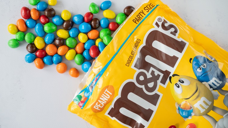 Close-up image of a bag of Peanut M&MS lying on a white surface with Peanut M&Ms pouring out of it