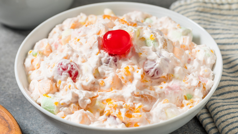 An Ambrosia salad with a cherry topping sits in a white bowl on a gray cabinet