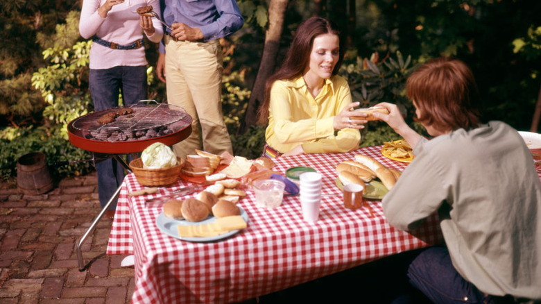 People stand around a grill while others eat at a picnic table in the 1970s