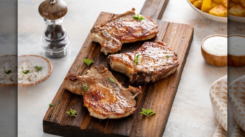 Three pork chops rest on a wooden cutting board by salt, pepper, and potatoes