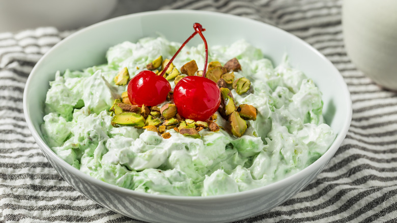 A bowl of Watergate salad topped with pistachios and cherries sits on a striped dishcloth