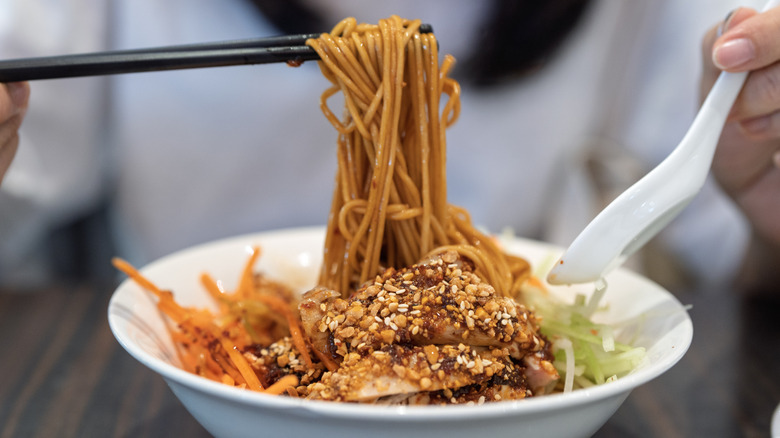 Hands using chopsticks to pull noodles form a bowl of chicken ramen with Szechuan chili pepper sauce