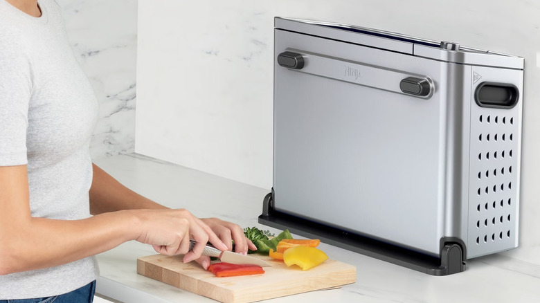 A Ninja Flip 10-in-1 Toaster Oven & Air Fryer with Added Broil Rack sits on a kitchen counter with vegetables in front of it.