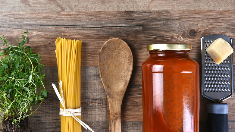 Herbs, spaghetti, a wooden spoon, a jar of sauce and parmesan on a wooden table