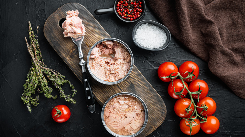 An opened can of tuna on a chopping board, with some fresh tomatoes, rock salt and dried thyme