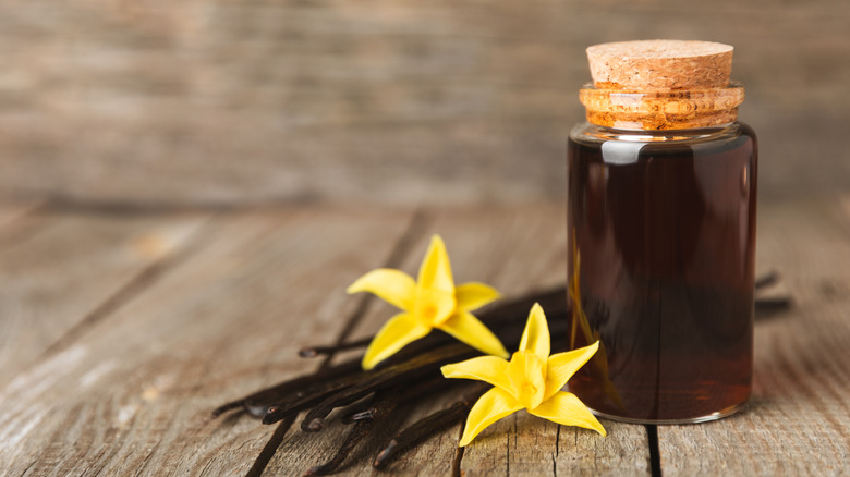 Vanilla pods and two yellow vanilla orchids sit beside a small jar of vanilla extract