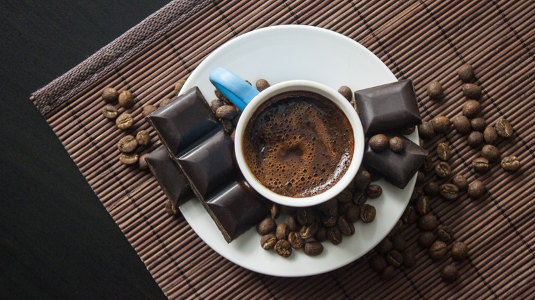 A cup of coffee sits on a white saucer with coffee beans and dark chocolate