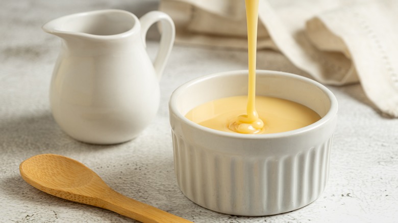 Condensed milk is poured into a white ramekin next to a wooden spoon and white creamer pot