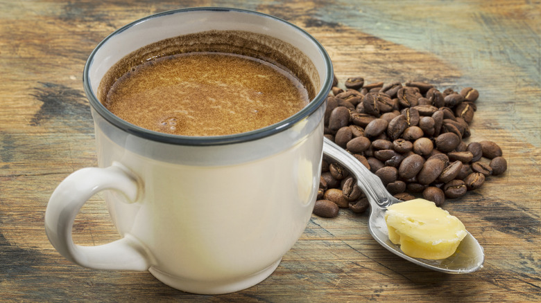 A mug of coffee sits beside a small pile of coffee beans and a spoon with a pat of butter on it on a wooden table