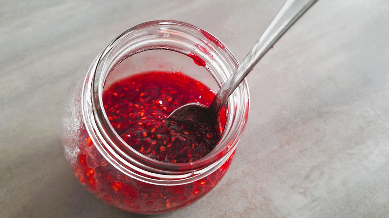Raspberry jam is in a half empty glass jar with a silver spoon