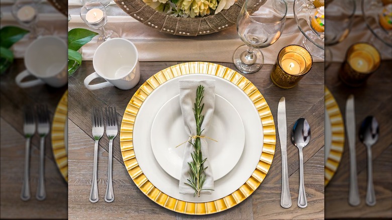 An elegantly set table with a gold-fluted charger plate beneath two other white plates, with a napkin on top tied with a sprig of rosemary