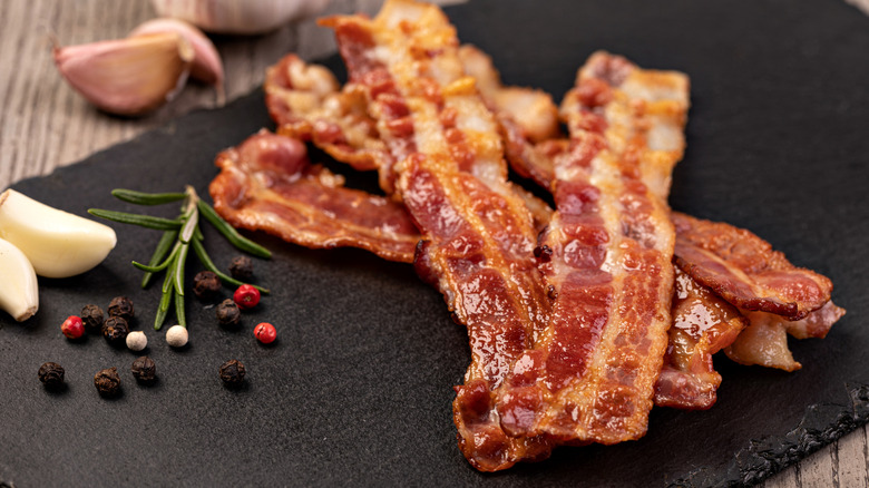 Close up of crispy bacon strips on a slate board with peppercorns, garlic cloves, and a rosemary sprig