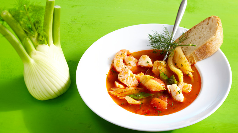 A bowl of hearty fish soup topped with fennel fronds sits next to a bulb of fennel.