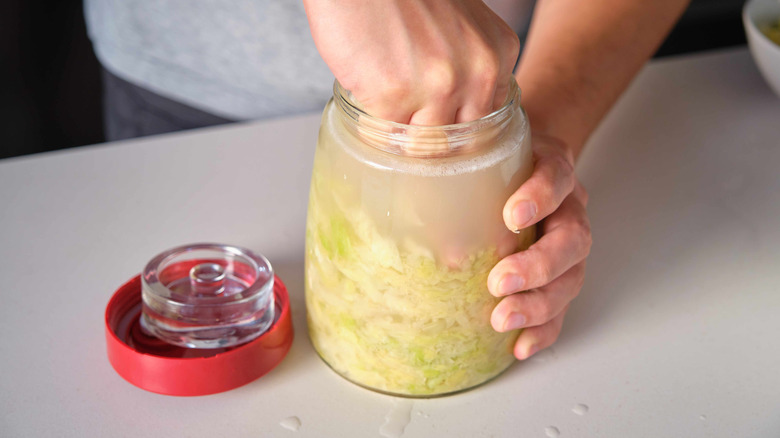 A hand pushing down sauerkraut in a jar, a weight and lid beside it