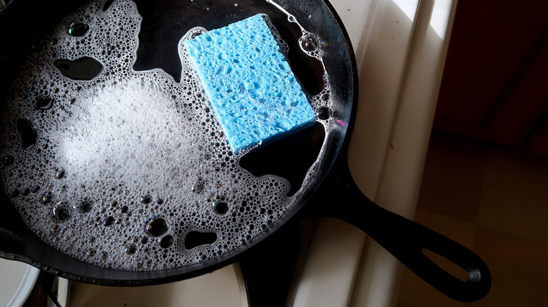 A cast-iron skillet is filled with soapy water and a blue sponge