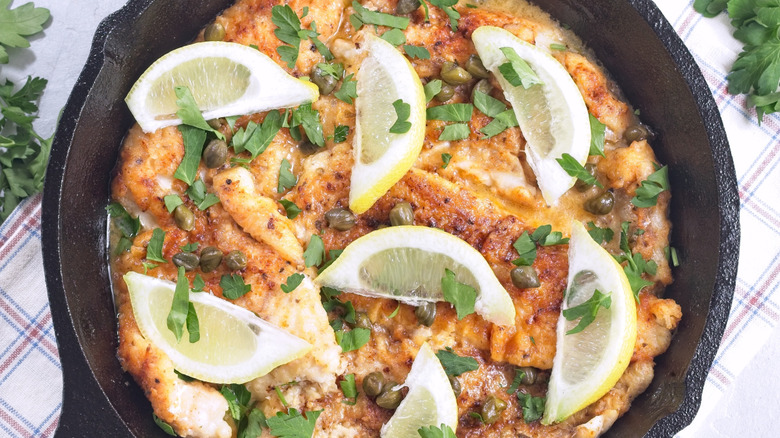 A cast-iron skillet holds chicken piccata with capers and lemon wedges