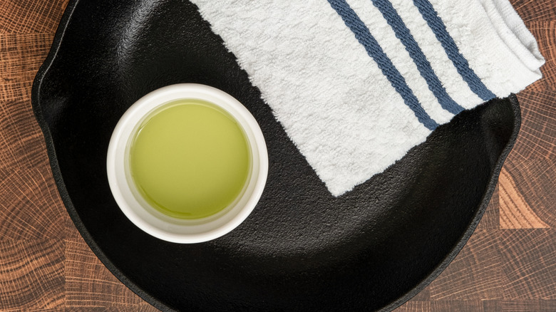 A ramekin of oil sits beside a white bar towel in a clean iron skillet