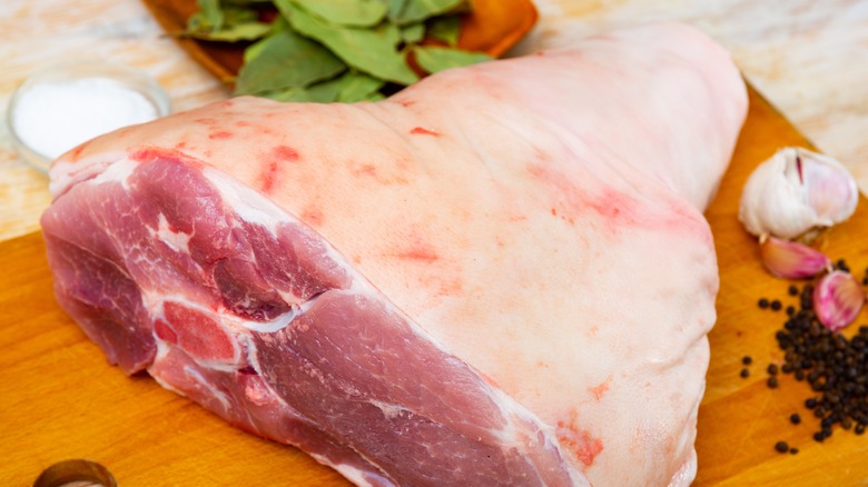 Raw pork leg on cutting board surrounded by fresh herbs and spices
