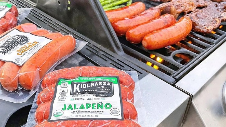 Packs of Kiolbassa sausage sit next to sausages cooking on a grill.