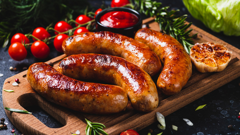 Grilled sausages sit on a wood board with ketchup, garlic, herbs, and tomatoes.