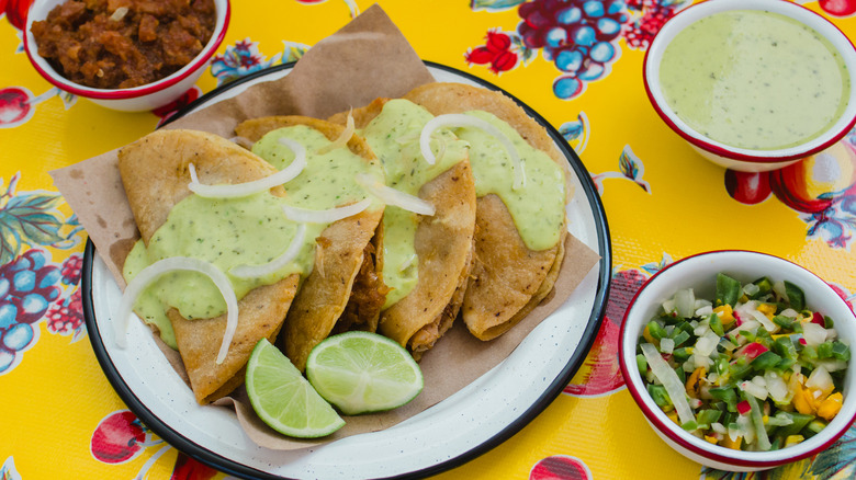 Four tacos with a green salsa poured over them on a bright yellow table cloth and two other side dishes