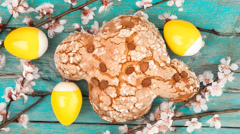 A traditional Italian Easter bread in the shape of a dove sits on a blue table with apple blossoms and dyed yellow eggs