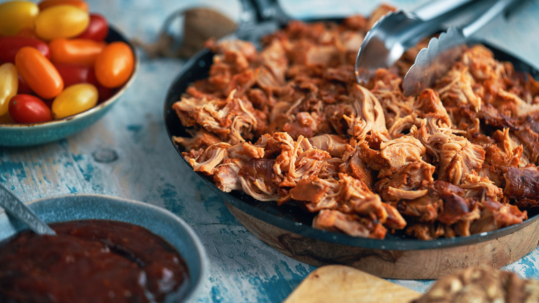 Close up of a pan of pulled buffalo chicken with tongs beside a bowl of ketchup and a bowl of grape tomatoes