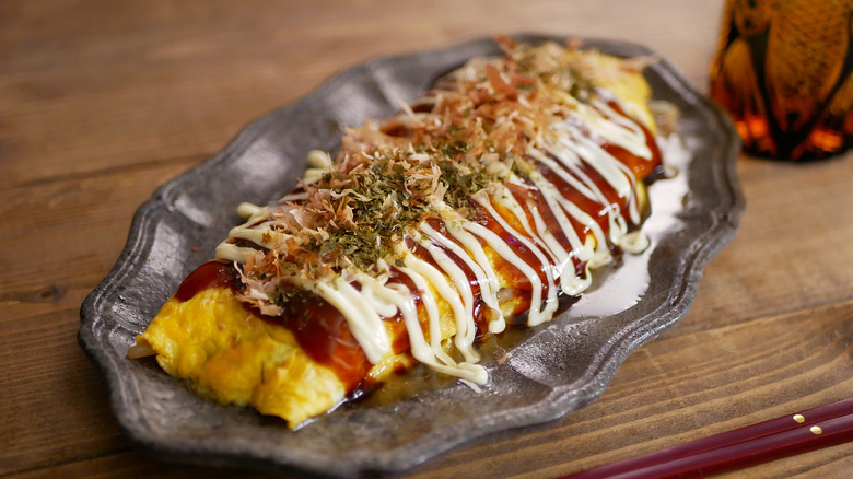 A Japanese omelet with pork belly and cabbage sits covered in sauces on a gray platter