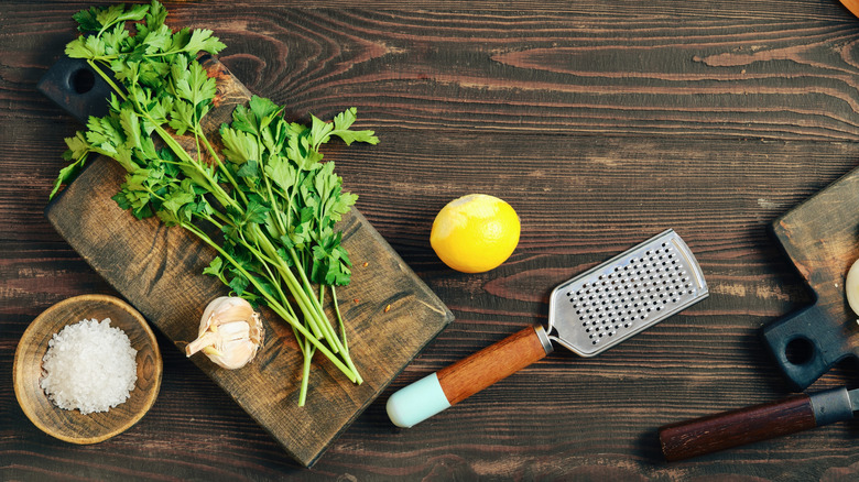 parsley on a wooden cutting board next to grater, lemon, garlic clove, and bowl of salt
