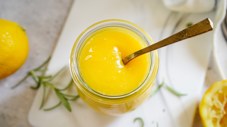 A jar of lemon curd with a spoon on a white cutting board with a rosemary sprig