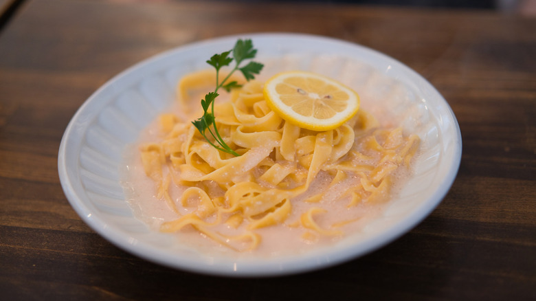 Pasta in a white bowl sits in a lemon sauce and is garnished with a lemon slice and a sprig of parsley