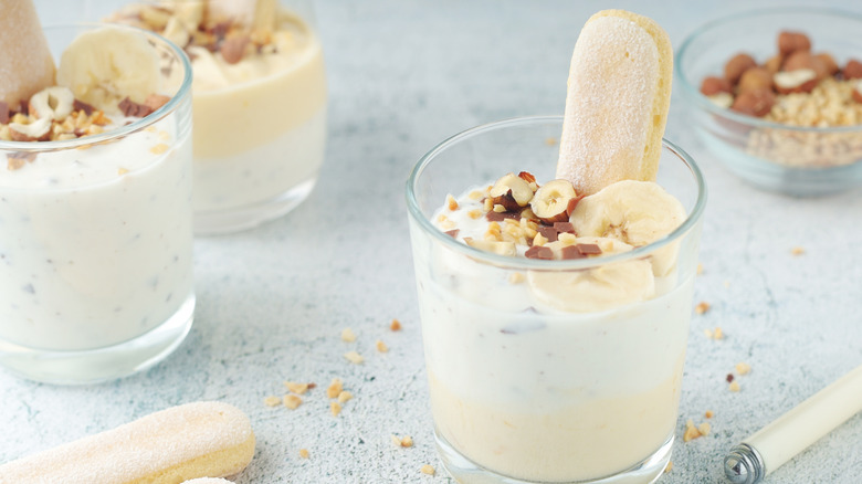 Individual yogurt puddings are topped with banana slices, hazelnuts, and a ladyfinger