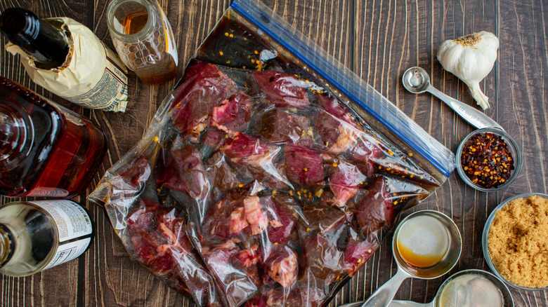 Chunks of red meat in a dark marinade in a plastic bag surrounded by spices and condiments