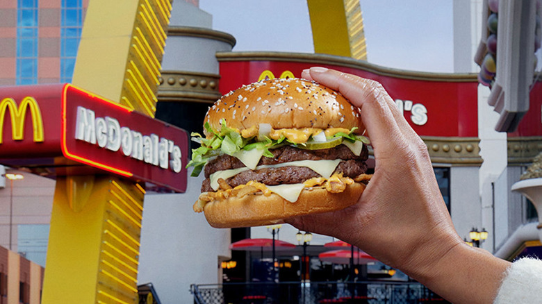 A hand holds out a Big Arch Burger in front of a McDonald's