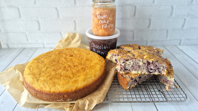 a yellow circle cake, slices of a swirled cake, a plastic bowl of chocolate frosting and a mason jar labeled peach cake filling