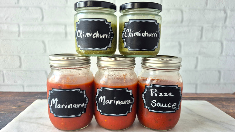 three mason jars in a row with tomato based sauces and two small jars of herb sauce on top