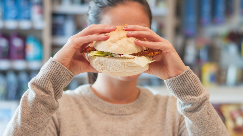A woman holds a sandwich in front of her face in front of a gas station beverage cooler.