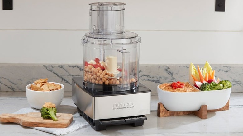 Cuisinart 14-cup food processor machine holding garbanzo beans on kitchen countertop next to a plated assortment of hummus, chips, and vegetables