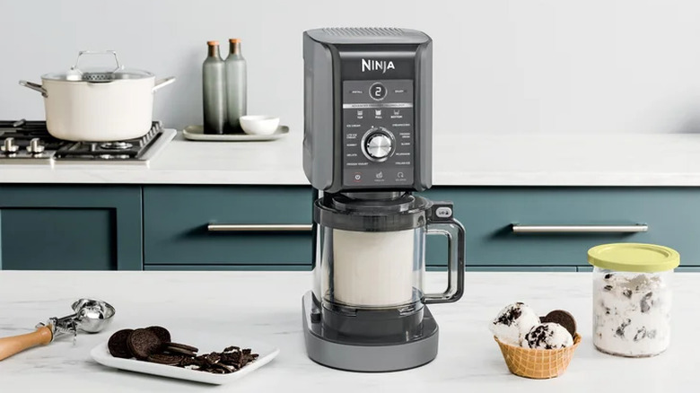 Ninja CREAMi Deluxe ice cream machine on countertop next to a pint and bowl of ice cream, assorted toppings, and an ice cream scooper