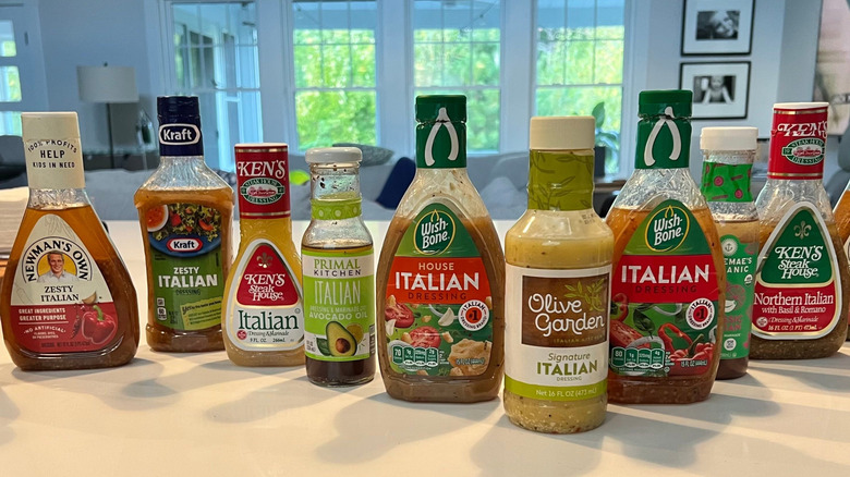 Eleven bottles of Italian Dressing stood in a row on a white kitchen island, from left to right: Ken's Steakhouse Italian, Newman's Own Family Recipe Italian, Ken's Steakhouse Northern Italian, Tessemae's Organic Italian, Wishbone Italian Dressing, Olive Garden Signature Italian, Wishbone House Italian, Primal Kitchen Italian, Kraft Zesty Italian, Newman's Own Zesty Italian, and Trader Joe's Organic Italian Dressing