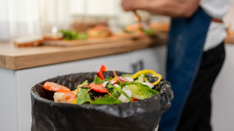 Garbage can containing good produce to show food waste, while an employee in an apron is in the background