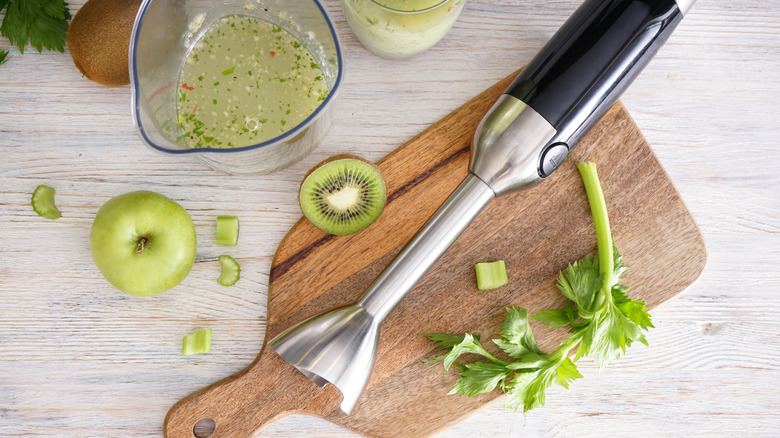 Immersion blender on wood cutting board with apples, kiwi, and celery