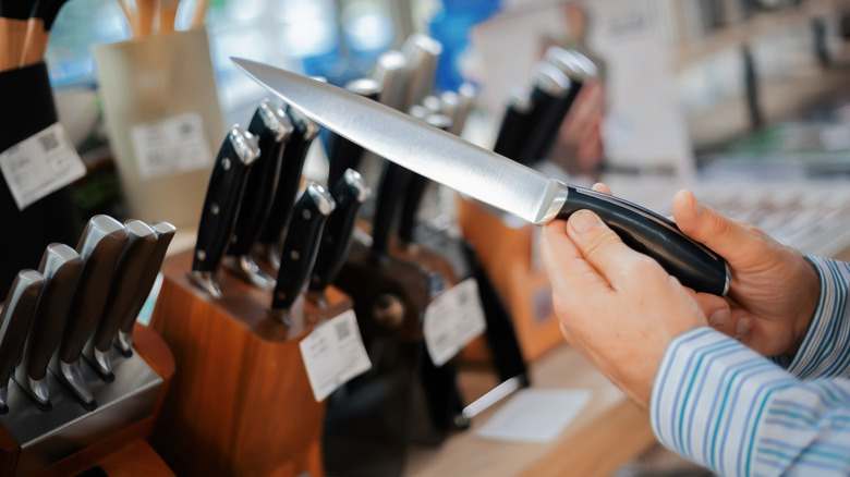 Close-up of hands holding a kitchen knife in a store, examining its blade and design among various knife sets on display