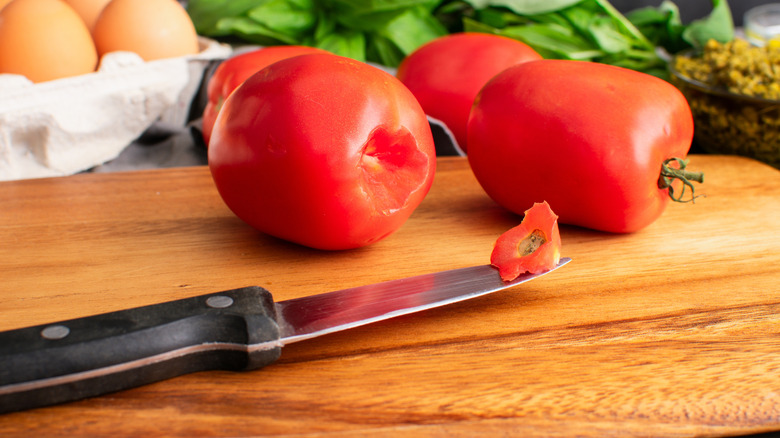 A paring knife sits on a cutting board with Roma tomatoes, and the knife has a core on its tip