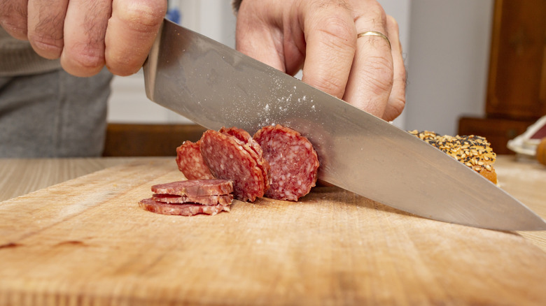 Hands use the belly of a chef's knife to slice salami on a wooden cutting board