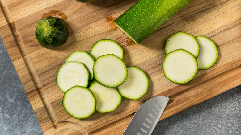 The tip of a chef's knife lays on a wooden cutting board surface with fresh slices of zucchini