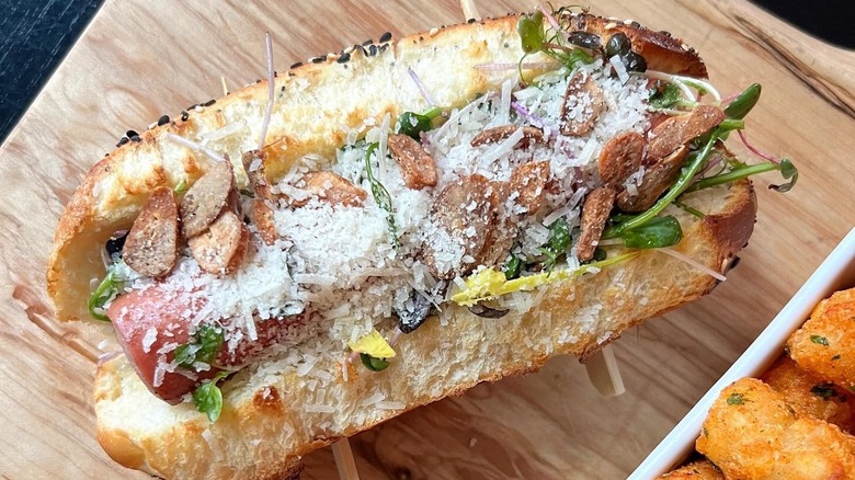 A beef hot dog topped with black truffle, garlic chips, greens, and grana padano cheese sits beside some tater tots