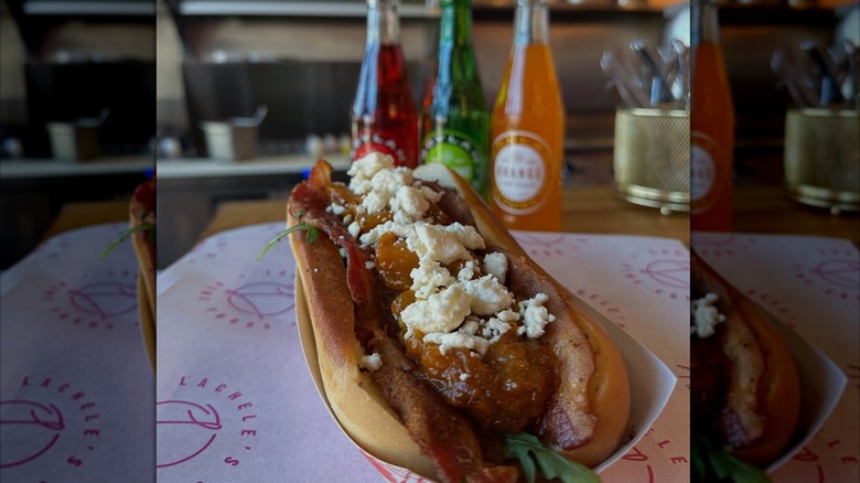 A hot dog is topped with bacon, crumbles of feta cheese, micro greens, and a generous helping of mango chutney