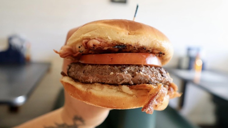 a hand holding a hamburger with bacon, tomato, and sauce on a bun